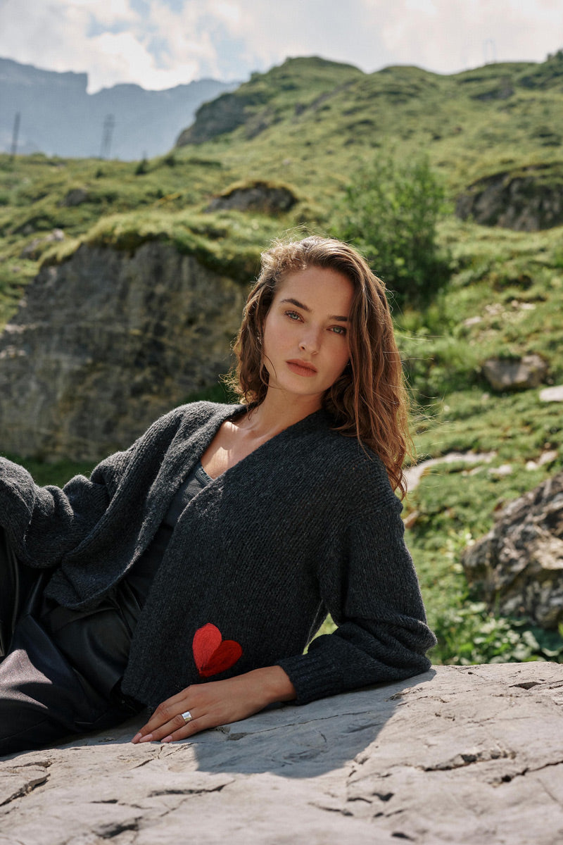Eine Frau mit welligem braunem Haar, die eine dunkle Strickjacke aus einer Merino-Alpaka-Mischung mit einem roten Herz-Intarsien-Aufnäher trägt, sitzt draußen auf einem Felsen. Mit ruhigem Gesichtsausdruck blickt sie in die Kamera. Im Hintergrund sind unter einem teilweise bewölkten Himmel grüne Hügel und felsige Klippen zu sehen.