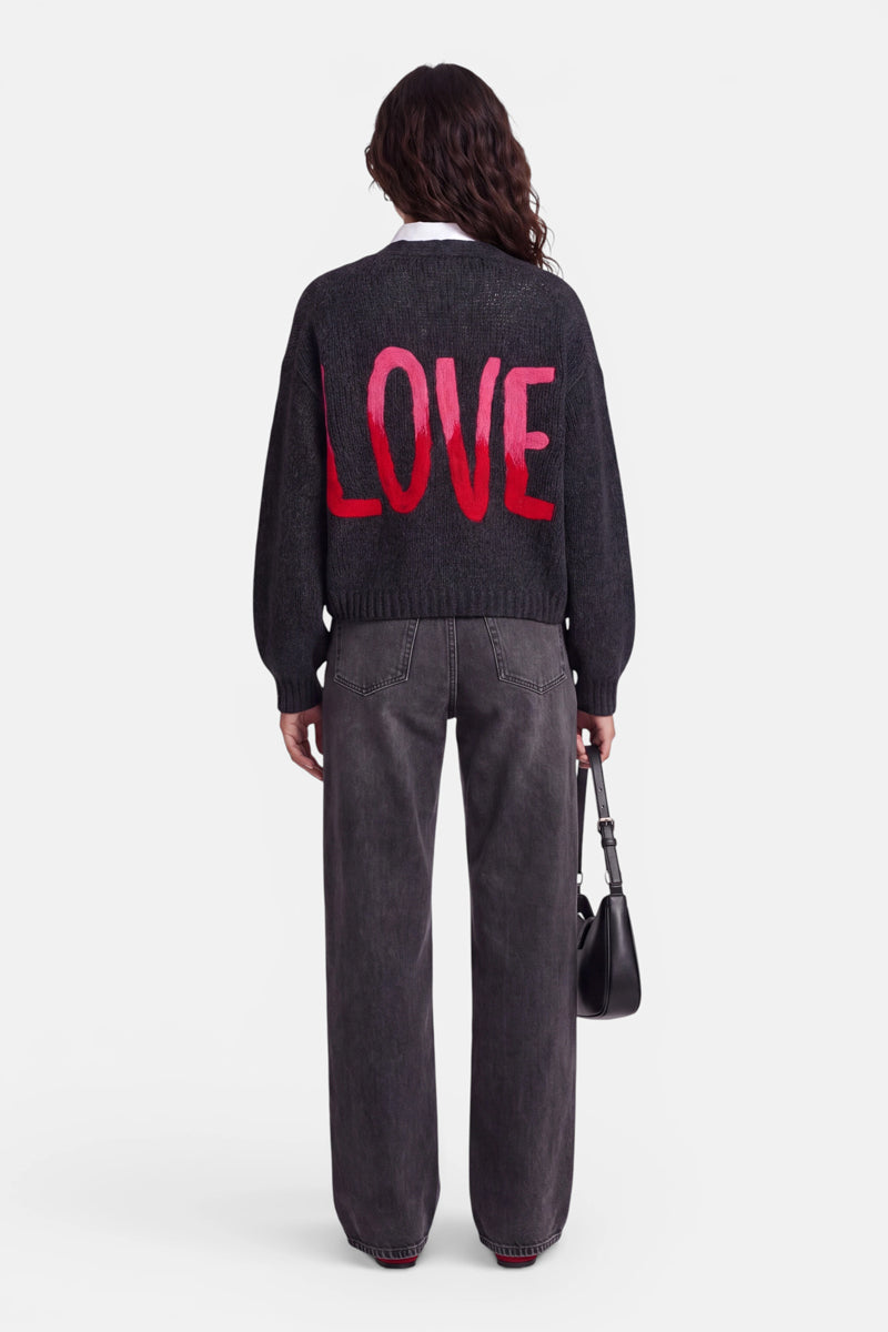 Eine Person mit welligem braunem Haar trägt eine dunkle Strickjacke aus einem kuscheligen Merino-Alpaka-Mix, auf deren Rücken in einer auffälligen Herz-Intarsie große rosa-rote Buchstaben das Wort „LOVE“ bilden. Dazu trägt sie eine weite graue Jeans und hält eine schwarze Handtasche in der Hand. Der Hintergrund ist schlicht weiß, und die Person ist von hinten zu sehen.