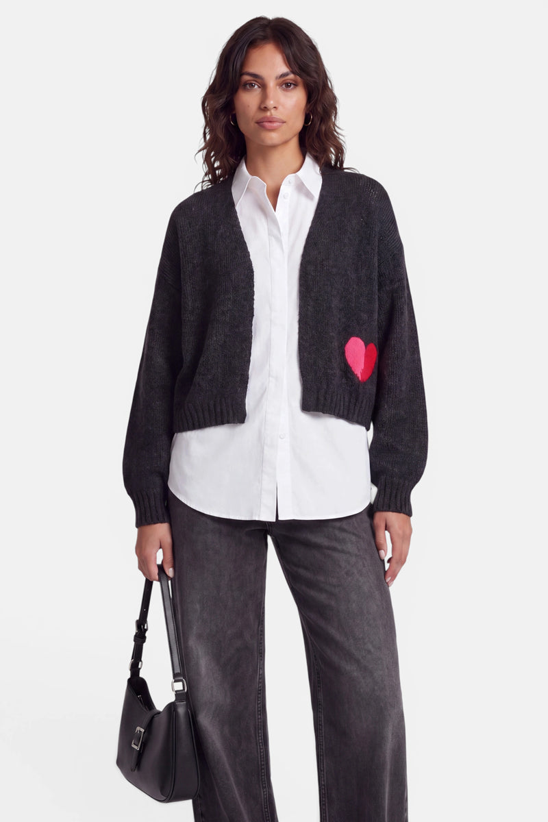 Eine Frau mit welligem braunem Haar trägt eine dunkelgraue Strickjacke aus Merino- und Alpaka-Wolle mit einer roten Herz-Intarsie auf einer Seite, darüber ein weißes Hemd mit Knöpfen und dazu eine lockere graue Jeans. Sie hält eine kleine schwarze Handtasche in der Hand und steht vor einem schlichten weißen Hintergrund, wobei sie direkt in die Kamera blickt.