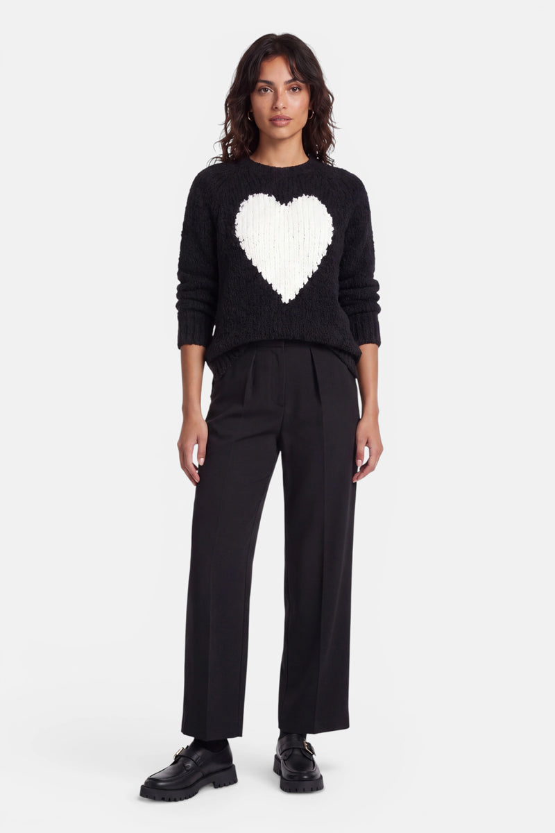 Eine Frau mit welligem, dunklem Haar trägt einen schwarzen Strickpullover aus Wollmischung mit einem großen weißen Herzmotiv auf der Vorderseite, dazu eine schwarze Hose mit weitem Bein und schwarze klobige Schuhe; sie steht vor einem schlichten weißen Hintergrund.