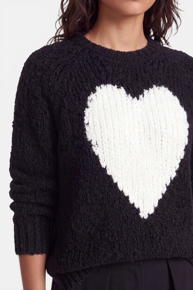Eine Frau mit welligem, dunklem Haar trägt einen strukturierten schwarzen Strickpullover mit einem großen, weißen Herzmotiv auf der Brust. Das Bild ist bis zu ihrer Nase und oberhalb ihrer Hüften beschnitten. Ihr linker Arm hängt entspannt an ihrer Seite. Der Hintergrund ist einfarbig weiß.