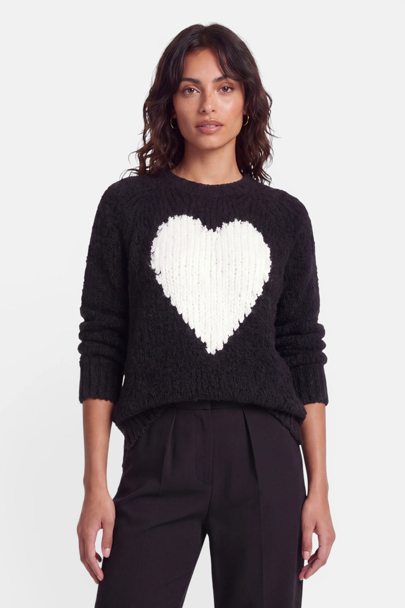 Eine Frau mit welligem, dunklem Haar trägt einen schwarzen Wollmix-Strickpullover mit einem großen weißen Herzmotiv auf der Brust. Sie steht mit dem Blick nach vorne, die Hände entspannt an den Seiten, und trägt eine schwarze Hose mit hoher Taille vor einem schlichten weißen Hintergrund.