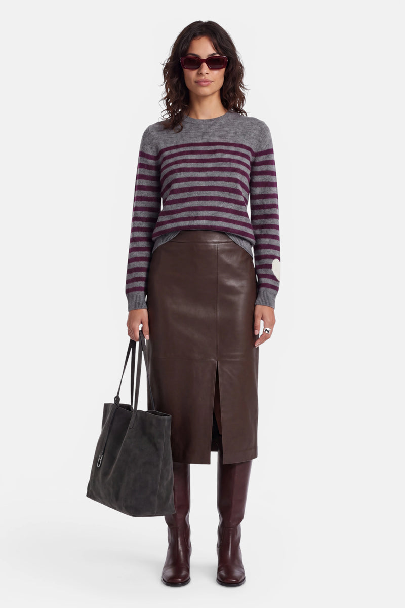 Eine Frau mit Sonnenbrille, einem grauen Strickpullover aus Alpaka-Wolle mit violetten Streifen und einem modischen Stehkragen, einem braunen Lederrock mit hoher Taille und einem Schlitz vorne, braunen kniehohen Stiefeln; sie hält eine große, dunkle Tragetasche in der Hand. Sie steht mit dem Blick nach vorne vor einem schlichten weißen Hintergrund.