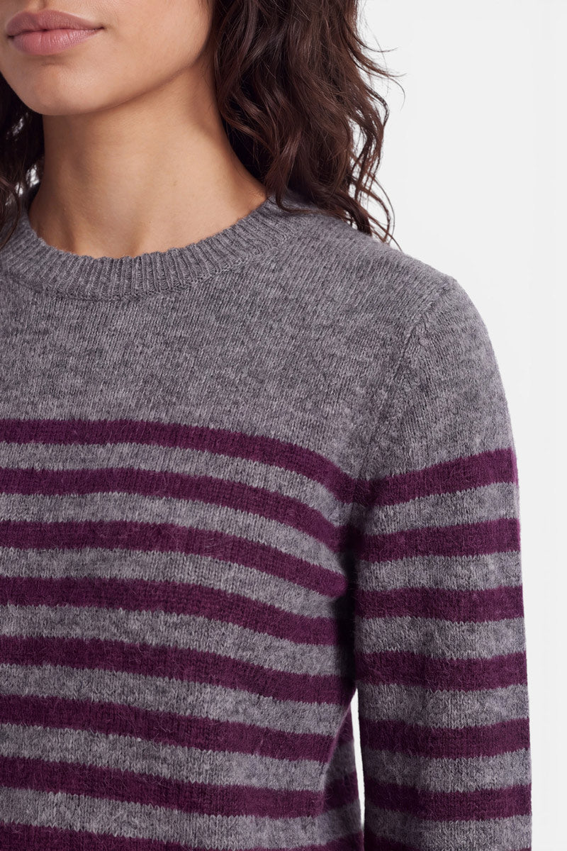 Eine Frau mit welligem braunem Haar trägt einen grauen Strickpullover aus Alpaka-Wolle mit horizontalen dunkelvioletten Streifen. Vor einem schlichten hellen Hintergrund sind nur ihr Gesicht (von der Nase abwärts) und ihr Oberkörper zu sehen. Der Pullover wirkt weich und dick.