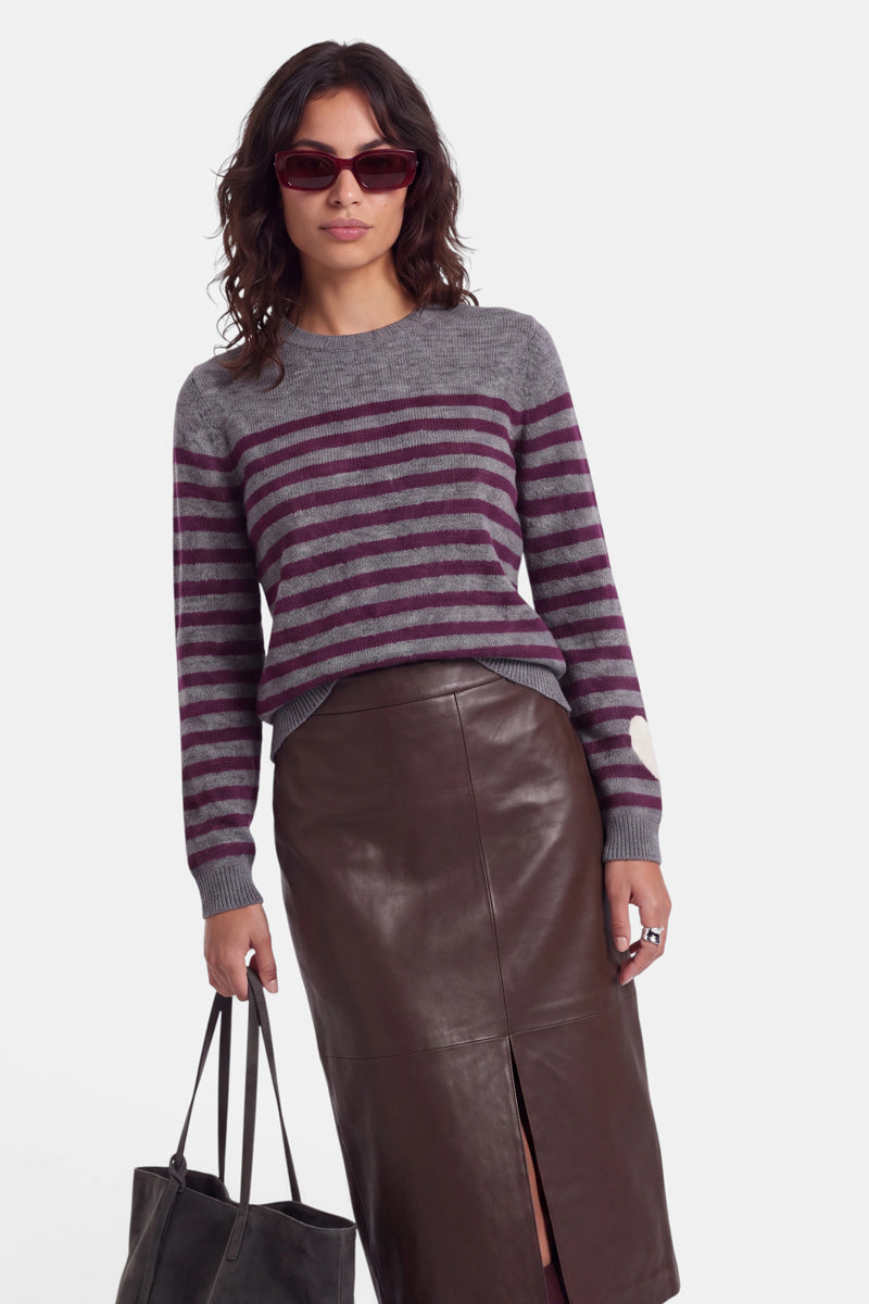 Eine Frau trägt eine Sonnenbrille und einen modischen Strickpullover für Damen – einen grau- und burgunderrot gestreiften Pullover mit einem Herz auf dem Ärmel –, dazu einen dunkelbraunen Lederrock mit einem Schlitz vorne. Sie trägt eine schwarze Tragetasche und hat welliges, schulterlanges dunkles Haar. Der Hintergrund ist schlicht weiß.