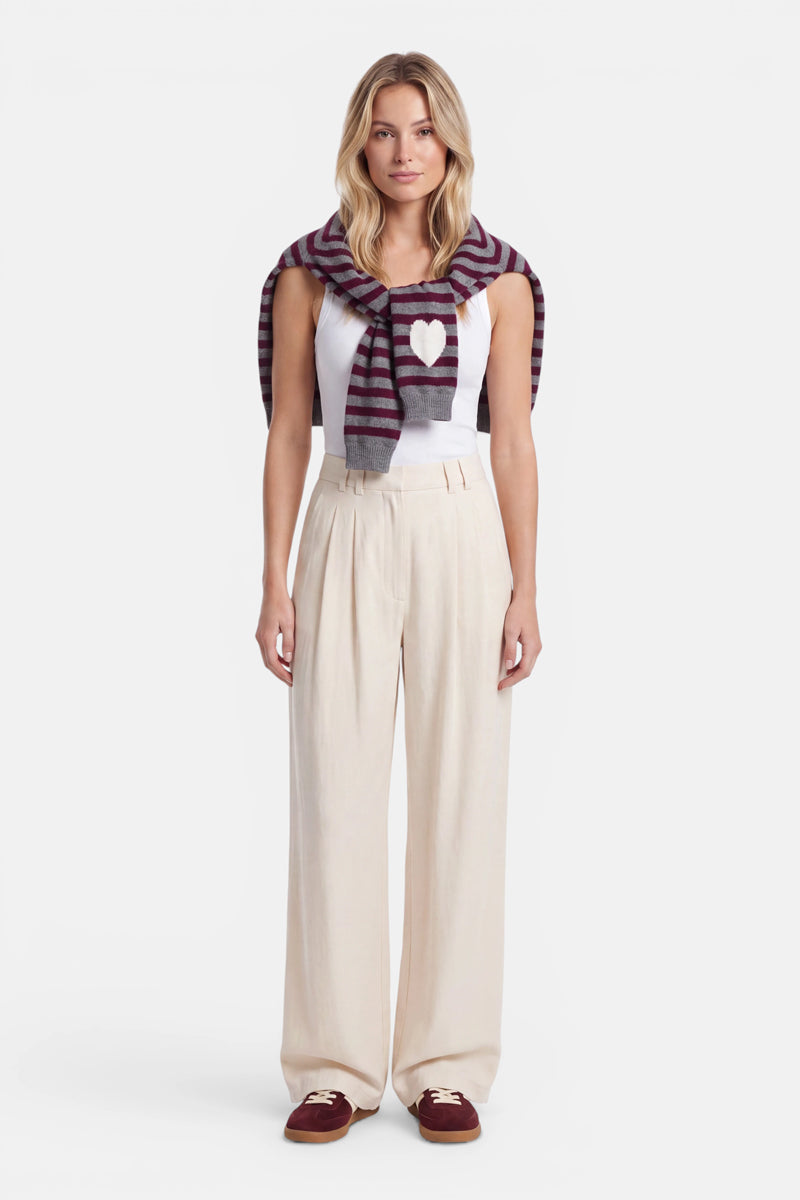 Vor einem schlichten weißen Hintergrund steht eine Frau mit dem Blick nach vorne. Sie trägt ein weißes Tanktop mit Herzmotiv, eine beige Hose mit hoher Taille und weitem Bein, burgunderrote Loafer sowie einen Strickpullover aus Alpaka-Wolle mit kastanienbraunen und grauen Streifen, der über ihre Schultern drapiert ist.