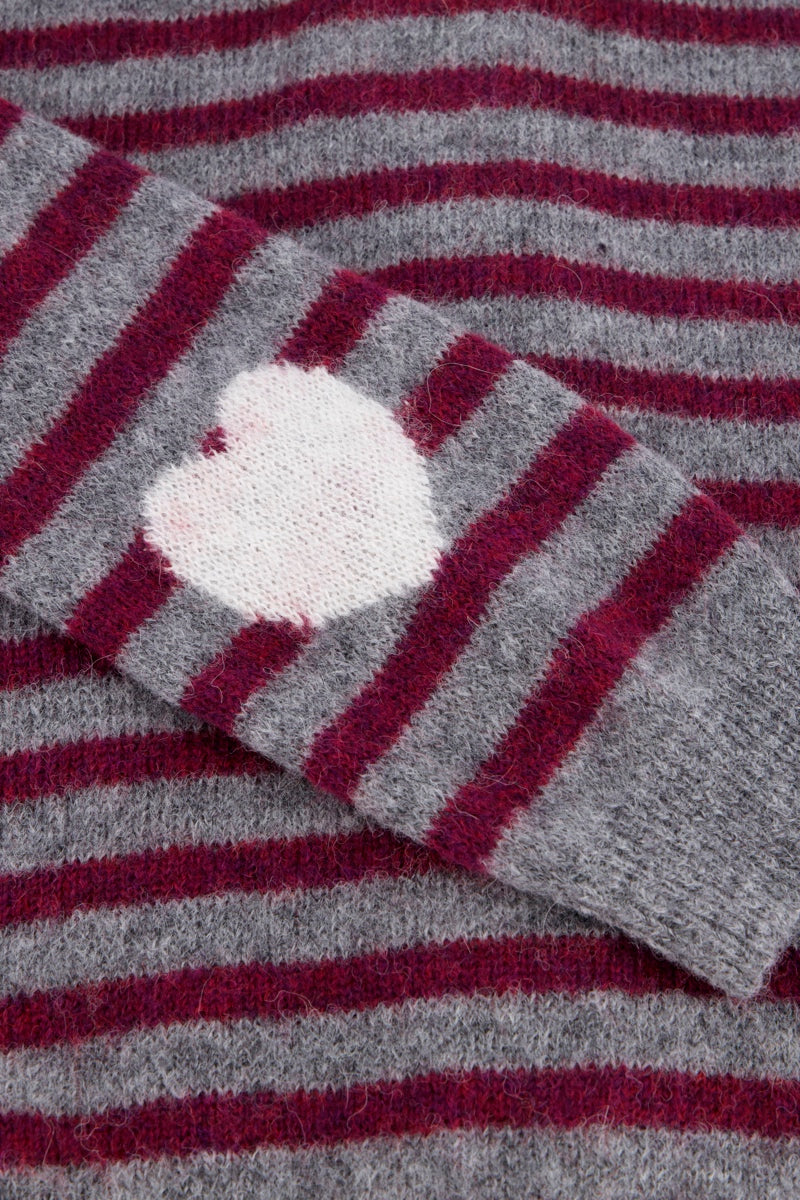 Eine Nahaufnahme eines grau- und burgunderrot gestreiften Wollpullovers („Streifen“) zeigt einen Ärmel mit einem weißen, herzförmigen Aufnäher („Herzmotiv“) am Ellbogen, der einen Kontrast zu den horizontalen Streifen sowohl am Ärmel als auch am Pulloverkörper bildet.