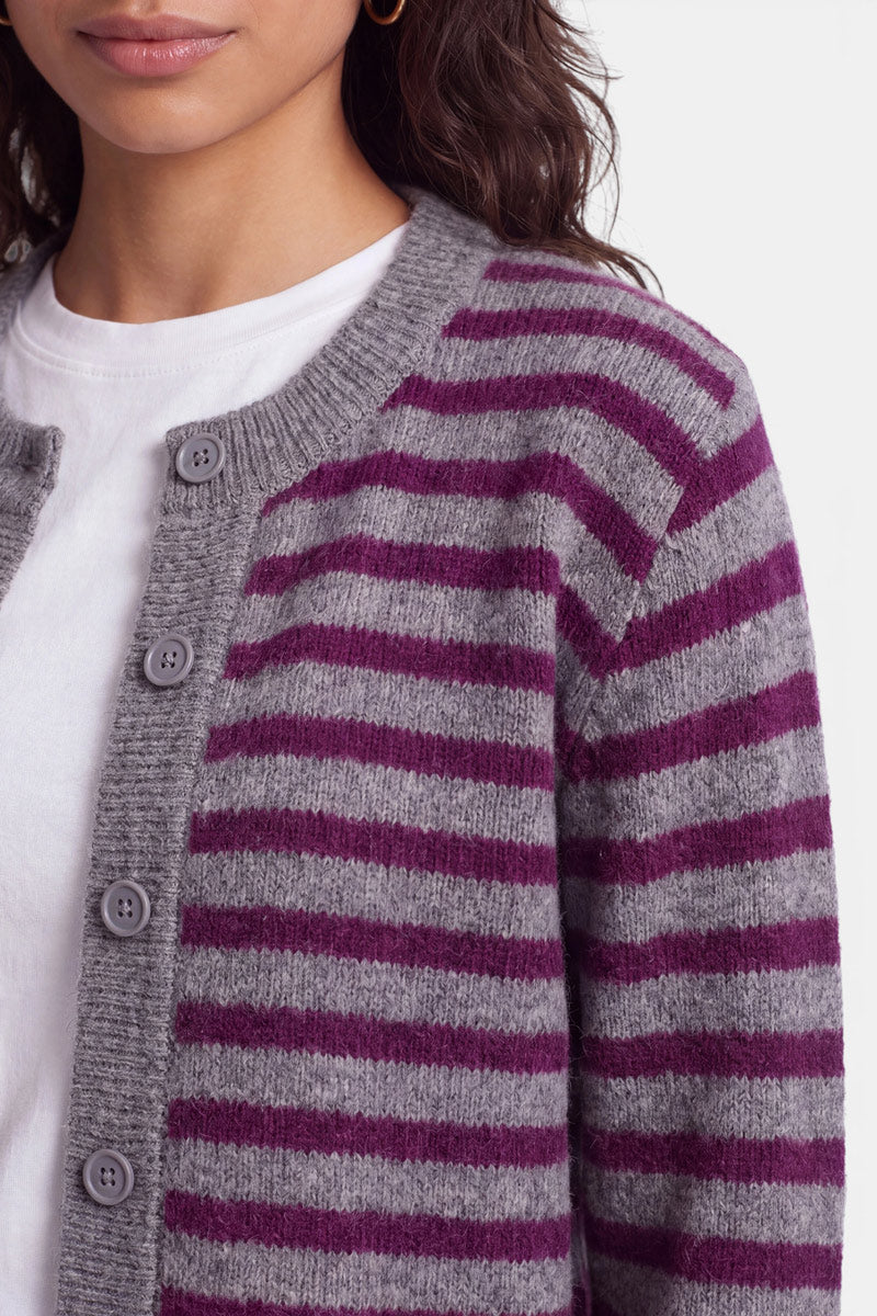 Eine Frau trägt eine graue Strickjacke aus Alpaka-Wolle mit horizontalen violetten Streifen über einem weißen T-Shirt. Die Strickjacke hat gerippte Säume und fünf graue Knöpfe. Vor einem schlichten weißen Hintergrund sind nur die untere Hälfte ihres Gesichts, ihr Hals und ihr Oberkörper zu sehen.