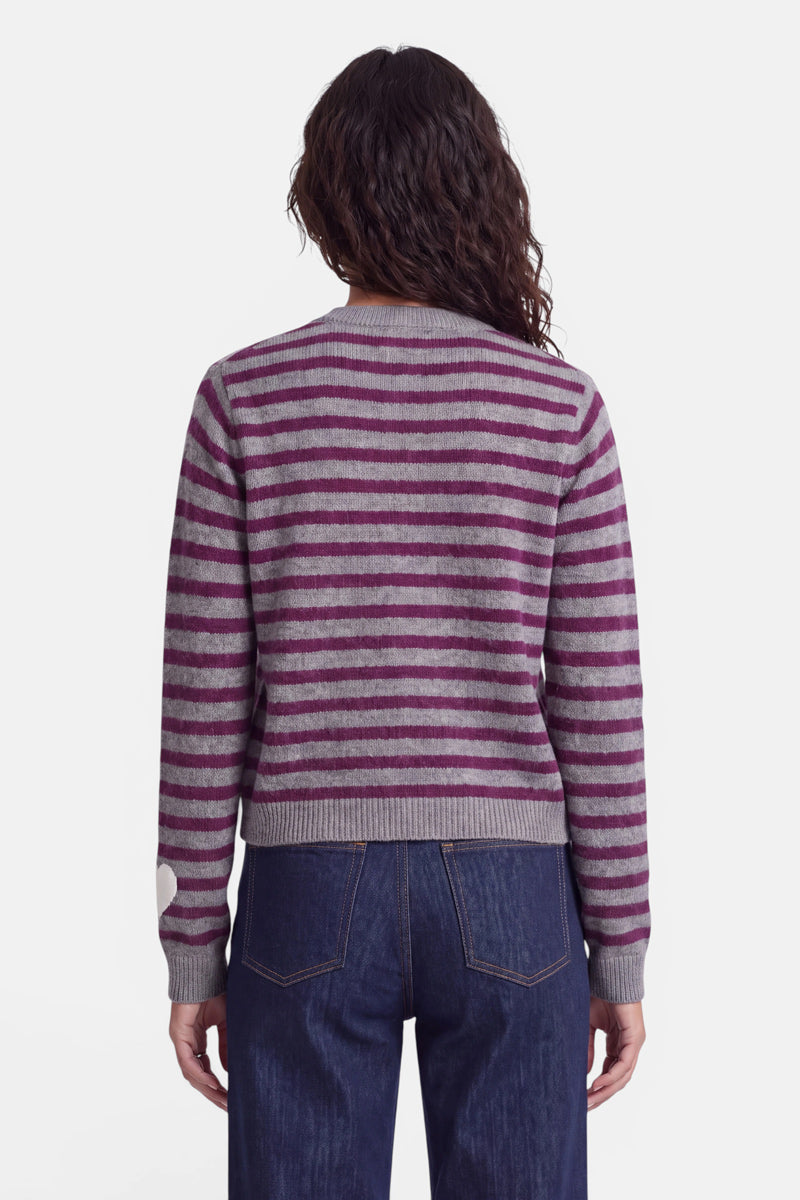 Eine Person mit welligem, dunklem Haar, von hinten gesehen, trägt eine graue Strickjacke aus Alpaka-Wolle mit horizontalen magentafarbenen Streifen und dunkelblaue Jeans. Die Strickjacke hat gerippte Bündchen und einen gerippten Saum und ist am linken Ärmel in der Nähe des Handgelenks mit einem kleinen weißen Herzmotiv verziert.