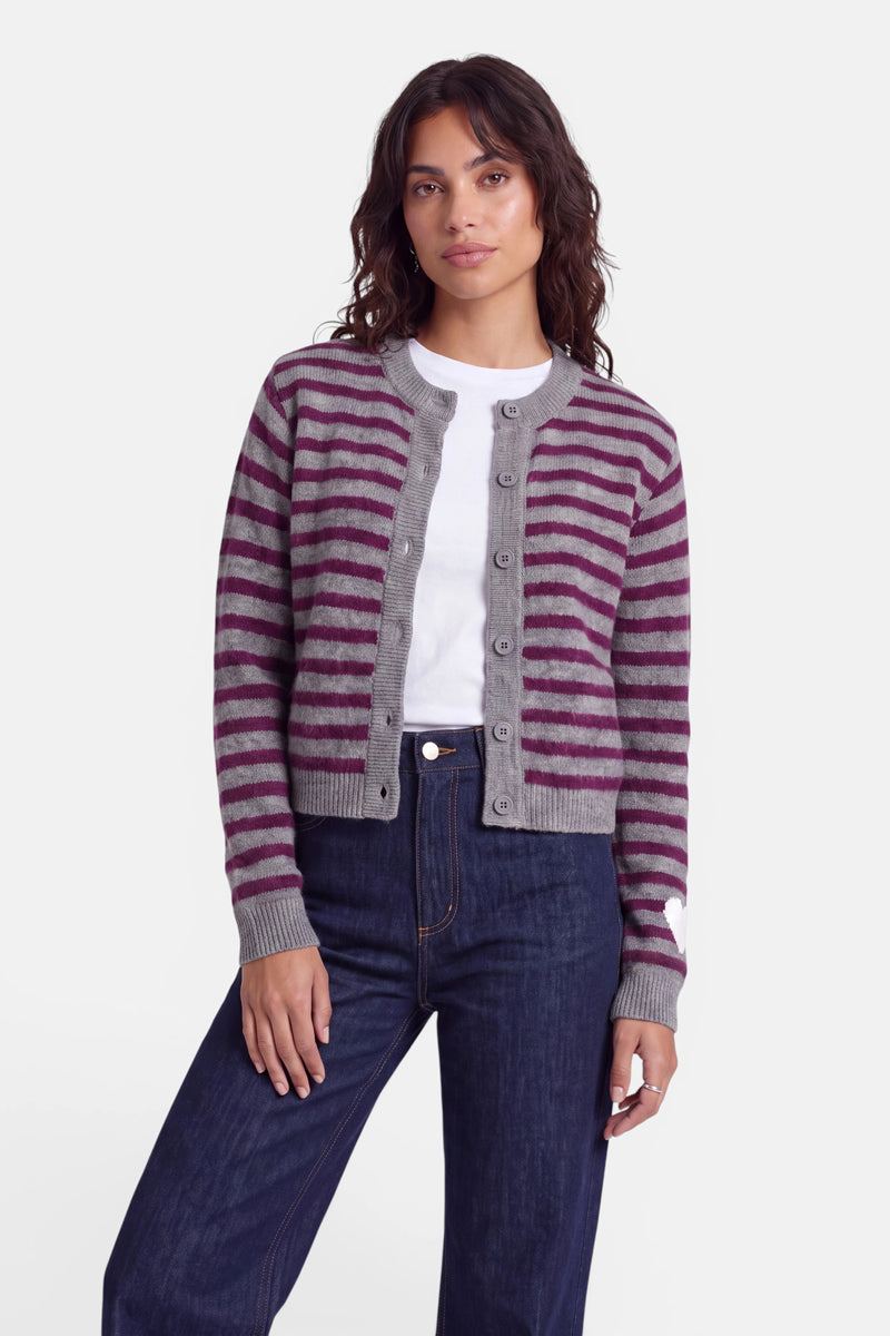 Eine Frau steht mit dem Blick nach vorne und trägt eine grau-lila gestreifte Strickjacke aus Alpaka-Wolle mit einem dezenten Herzmotiv über einem weißen T-Shirt und dunkelblauen Jeans. Sie hat mittellanges, welliges Haar und einen neutralen Gesichtsausdruck. Der Hintergrund ist schlicht weiß.