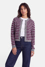 Eine Frau steht mit dem Blick nach vorne und trägt eine grau-lila gestreifte Strickjacke aus Alpaka-Wolle mit einem dezenten Herzmotiv über einem weißen T-Shirt und dunkelblauen Jeans. Sie hat mittellanges, welliges Haar und einen neutralen Gesichtsausdruck. Der Hintergrund ist schlicht weiß.