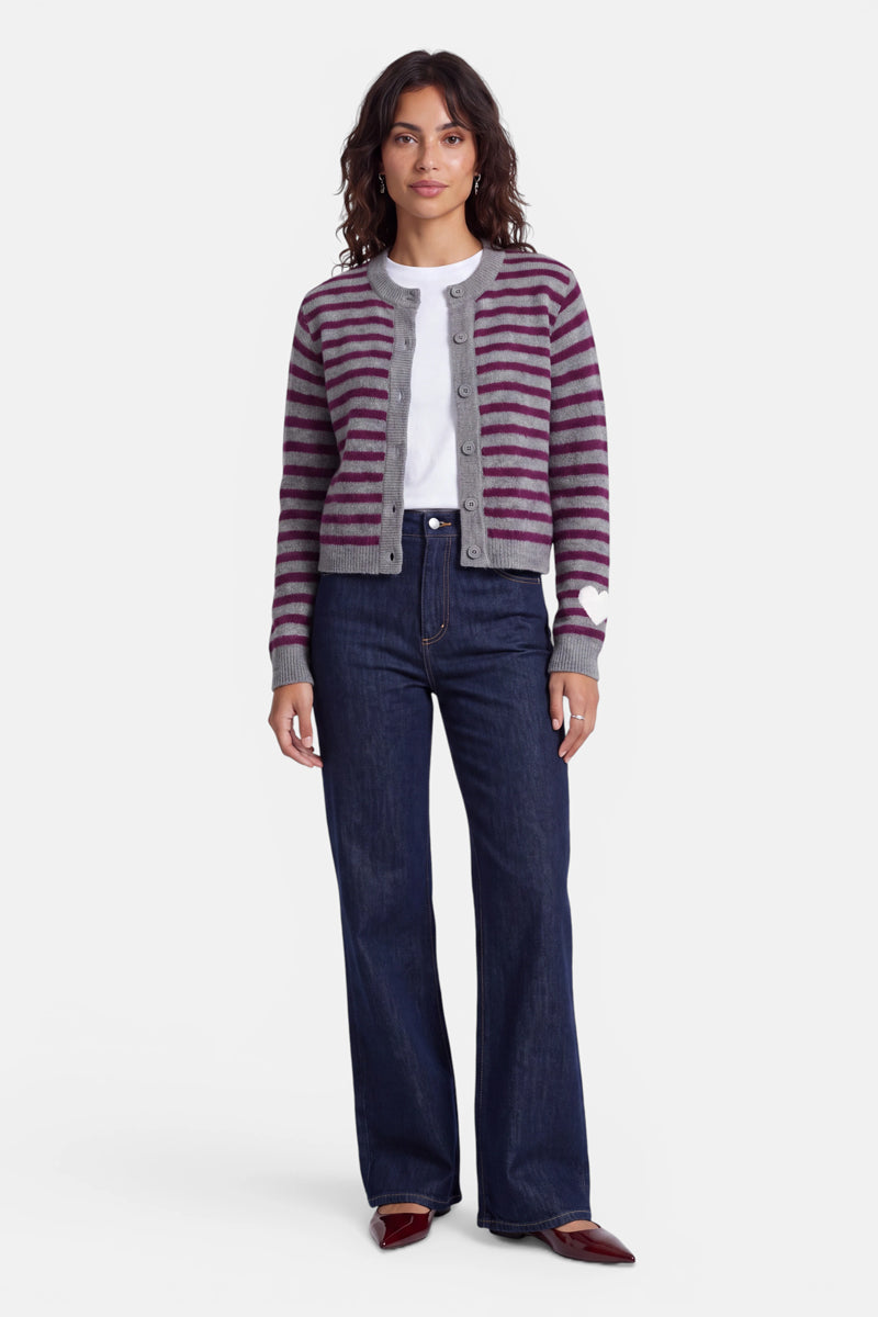 Eine Frau mit welligem braunem Haar trägt eine grau-lila gestreifte Strickjacke aus Alpaka-Wolle über einem weißen T-Shirt, dunkelblauen Jeans mit hoher Taille und burgunderroten spitzen Schuhen. Sie steht mit neutralem Gesichtsausdruck und entspannt an den Seiten hängenden Armen vor einem einfarbigen Hintergrund und blickt geradeaus.