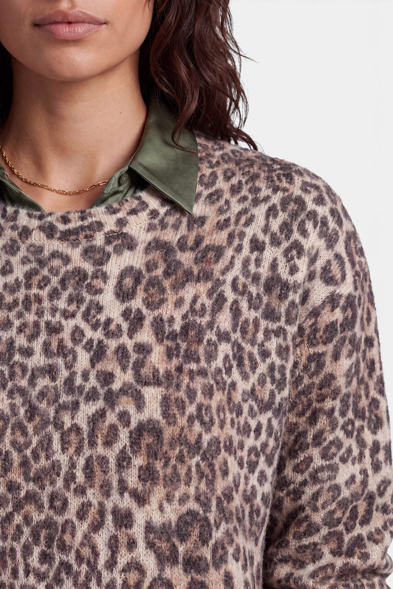 Eine Frau trägt einen flauschigen, übergroßen Strickpullover mit Leopardenmuster aus einer luxuriösen Woll-Alpaka-Mischung über einem glänzenden, olivgrünen Hemd mit Kragen und einer goldenen Halskette. Sie hat welliges braunes Haar und trägt dezentes Make-up. Das Bild ist von ihrer Brust bis zur Gesichtsmitte beschnitten und hat einen schlichten weißen Hintergrund.