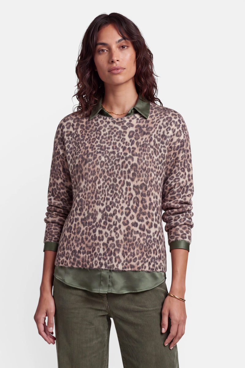 Eine Frau mit welligem braunem Haar trägt einen übergroßen Strickpullover mit Leopardenmuster aus einer Woll-Alpaka-Mischung über einem olivgrünen Hemd mit Kragen und einer dazu passenden Cordhose. Sie steht mit neutralem Gesichtsausdruck vor einem schlichten weißen Hintergrund und blickt geradeaus.