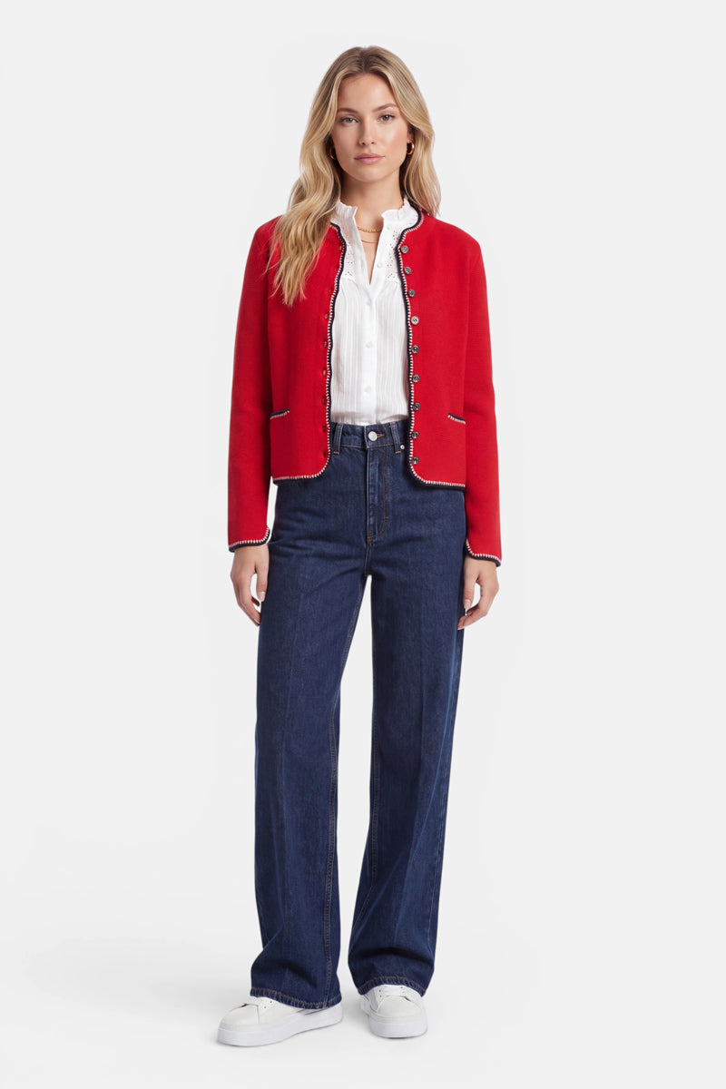 Eine Frau steht mit dem Blick nach vorne und trägt eine leuchtend rote Strickjacke im bayerischen Stil – einen bezaubernden Damen-Cardigan aus einer luxuriösen Wolle-Kaschmir-Mischung – über einer weißen Bluse, dazu dunkelblaue Jeans mit hoher Taille und weitem Bein sowie weiße Sneaker. Ihr langes blondes Haar ist in lockeren Wellen gestylt, während sie vor einem schlichten weißen Hintergrund posiert.
