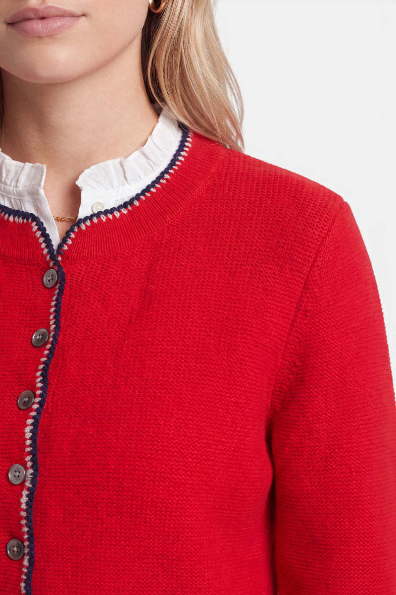 Eine Frau trägt über einer weißen Rüschenbluse eine strukturierte rote Damen-Strickjacke aus einem luxuriösen Wolle-Kaschmir-Mix mit schwarzen Knöpfen und marineblauen Besätzen. Zu sehen sind nur ihr unterer Gesichtsbereich, ihr Hals und ein Teil ihrer Schulter. Sie hat blondes Haar und trägt einen kleinen goldenen Creolen-Ohrring.