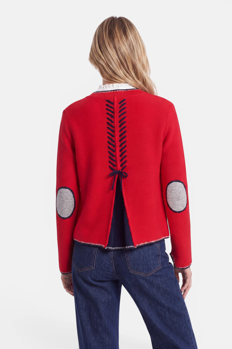 Eine Person mit langen blonden Haaren trägt eine Damen-Strickjacke in einem satten Rotton mit marineblauen Schnürdetails am Rücken, einem marineblauen Seitenschlitz, grauen Ellbogenpatches und kontrastierenden Bündchen. Diese Strickjacke im bayerischen Stil ist aus einer luxuriösen Wolle-Kaschmir-Mischung gefertigt und wird zu einer blauen Jeans getragen; die Person wird von hinten vor einem schlichten weißen Hintergrund fotografiert.