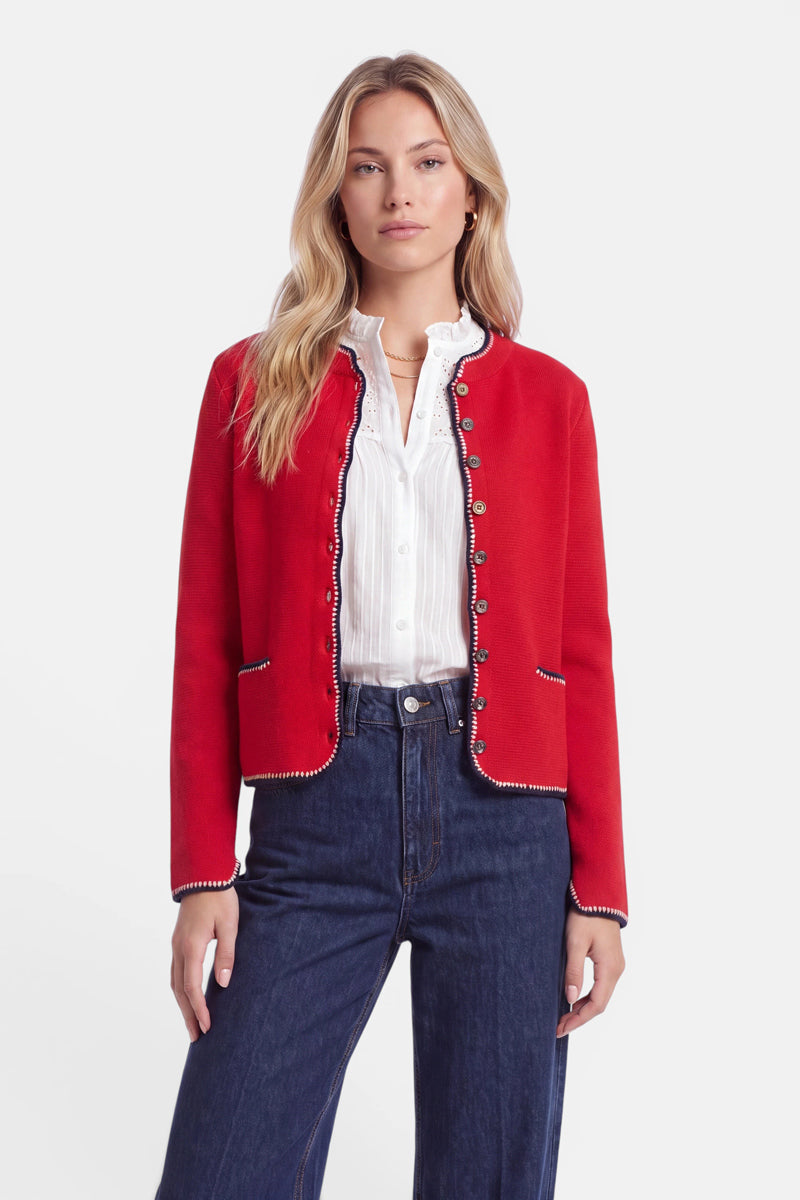Eine Frau steht mit dem Gesicht nach vorne und trägt eine rote Strickjacke im bayerischen Stil mit silbernen Knöpfen und schwarzer Borte über einer weißen Rüschenbluse und einer blauen Jeans mit hoher Taille. Sie hat langes blondes Haar und einen ausdruckslosen Gesichtsausdruck, vor einem schlichten weißen Hintergrund.