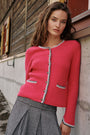 Eine Frau mit welligem braunem Haar trägt eine leuchtend rosa Strickjacke im bayerischen Stil aus einer Woll-Kaschmir-Mischung mit grauen Besätzen, silbernen Knöpfen und zarter Hirsch-Stickerei. Dazu trägt sie eine graue Plisseehose und lehnt an einer rustikalen Holzwand mit einem kleinen Fenster, während sie mit ausdruckslosem Blick in die Kamera schaut.