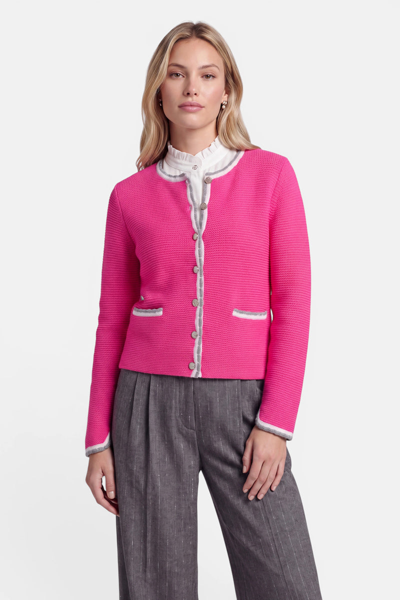 Eine Frau mit langen blonden Haaren trägt eine leuchtend rosa Strickjacke im bayerischen Stil mit weißem Rand über einer weißen Rüschenbluse und einer grauen Nadelstreifenhose. Die aus einem luxuriösen Wolle-Kaschmir-Mix gefertigte, geknöpfte Strickjacke ist auf der Vorderseite mit dezenten Hirsch-Stickereien verziert. Sie steht mit neutralem Gesichtsausdruck vor einem schlichten weißen Hintergrund und blickt nach vorne.