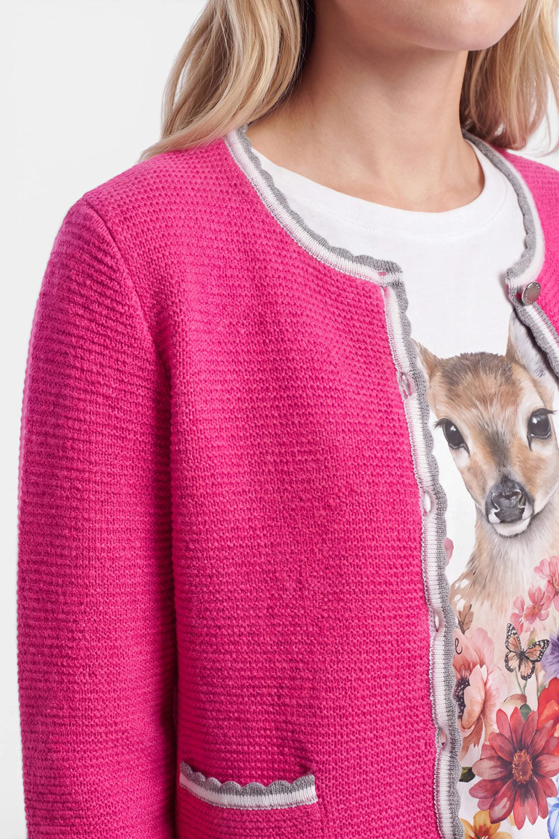 Eine Frau trägt eine strukturierte, leuchtend rosa Strickjacke im bayerischen Stil mit grauer Borte über einem weißen T-Shirt, auf dem ein realistisch dargestelltes Reh inmitten von rosa Blumen abgebildet ist. Nur ihr unterer Gesichtsbereich und ihr Oberkörper sind zu sehen; ihr Haar ist blond und wellig. Der Wolle-Kaschmir-Mix der Strickjacke verleiht dem Outfit sowohl Weichheit als auch Wärme.