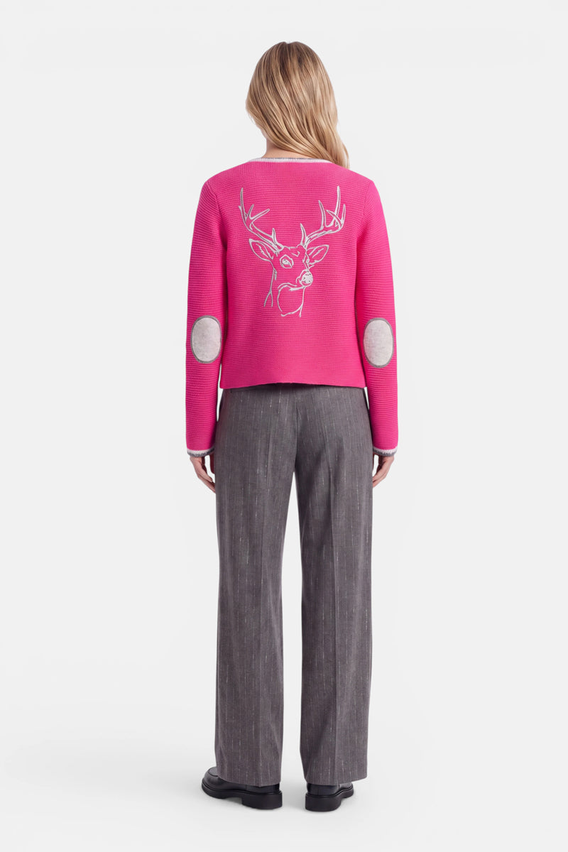 Eine Person steht mit dem Rücken zur Kamera und trägt eine leuchtend pinkfarbene Strickjacke im bayerischen Stil aus einer luxuriösen Wolle-Kaschmir-Mischung, auf deren Rücken eine weiße Hirsch-Stickerei (gestickter Hirschkopf) zu sehen ist. Die Strickjacke hat graue, ovale Ellbogenflicken und wird zu einer grauen Hose mit weitem Bein und dezenten Streifen sowie schwarzen Schuhen getragen. Der Hintergrund ist schlicht weiß. Die Person hat langes Haar.