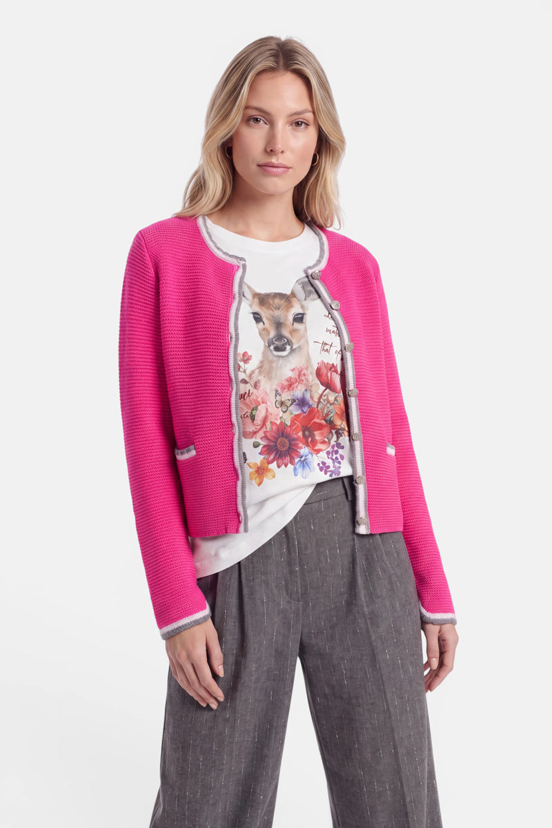Eine Frau mit offenem blondem Haar trägt eine leuchtend rosa Strickjacke im bayerischen Stil mit grauer Borte aus einem luxuriösen Wolle-Kaschmir-Mix über einem weißen T-Shirt mit Blumen- und Tierprint. Dazu trägt sie eine graue Nadelstreifenhose und steht vor einem schlichten weißen Hintergrund, während sie selbstbewusst in die Kamera blickt. Dezente Hirsch-Stickereien auf der Strickjacke verleihen ihr eine elegante Note.