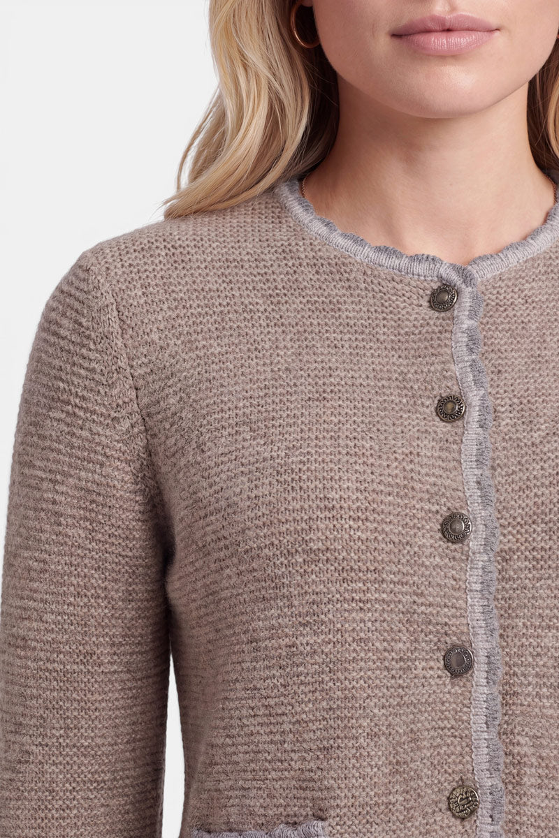 Eine Frau mit hellem, gewelltem Haar trägt eine strukturierte, beige Strickjacke im bayerischen Stil mit runden Silberknöpfen und hellgrauer, gewellter Borte. Das Bild ist von ihren Schultern bis knapp über ihre Taille beschnitten, und ihre Lippen sind zu einem neutralen Gesichtsausdruck geschlossen.