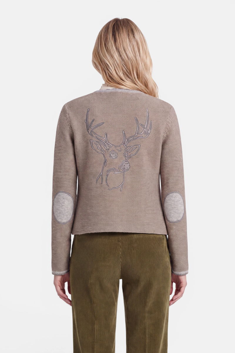 Eine Frau mit welligem blondem Haar, von hinten gesehen, trägt eine beige Strickjacke im bayerischen Stil aus einem weichen Woll-Kaschmir-Mix. Die Strickjacke ist auf der Rückseite mit einer gestickten Hirschkopf-Kontur – Hirschstickerei – verziert und hat graue Ellbogenpatches. Dazu trägt sie eine olivgrüne Cordhose. Der Hintergrund ist schlicht weiß.