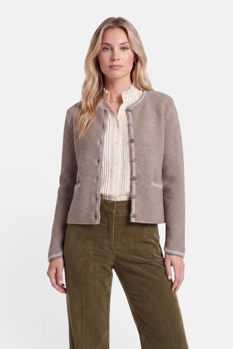 Eine Frau steht mit dem Blick nach vorne und trägt eine taupefarbene, strukturierte Strickjacke im bayerischen Stil mit runden Knöpfen, eine cremefarbene Bluse mit Rüschen und eine olivgrüne Cordhose. Sie hat langes, welliges blondes Haar und einen neutralen Gesichtsausdruck. Der Hintergrund ist schlicht weiß.