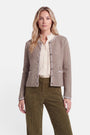 Eine Frau steht mit dem Blick nach vorne und trägt eine taupefarbene, strukturierte Strickjacke im bayerischen Stil mit runden Knöpfen, eine cremefarbene Bluse mit Rüschen und eine olivgrüne Cordhose. Sie hat langes, welliges blondes Haar und einen neutralen Gesichtsausdruck. Der Hintergrund ist schlicht weiß.