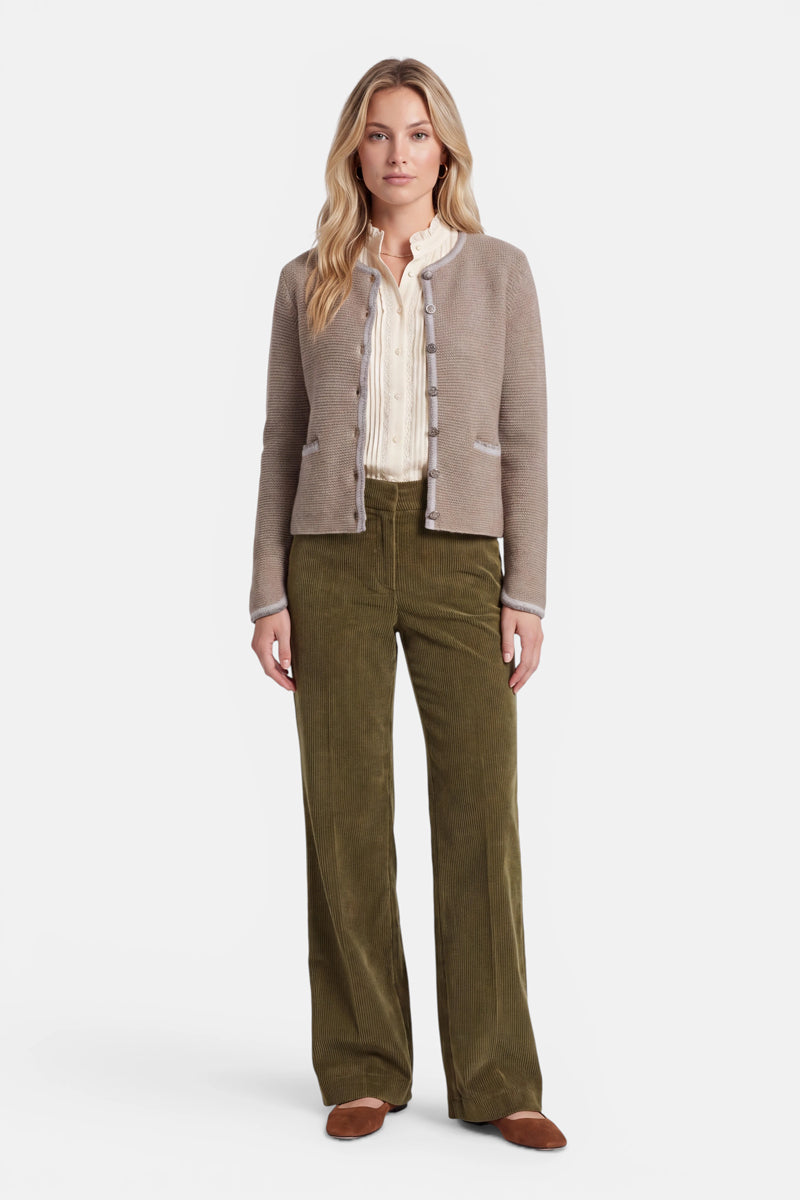 Vor einem weißen Hintergrund steht eine Frau, die eine beige Strickjacke im bayerischen Stil mit Zierknöpfen, eine cremefarbene Rüschenbluse, eine olivgrüne Cordhose mit weitem Bein und braune Schuhe trägt. Die Strickjacke ist aus einer luxuriösen Woll-Kaschmir-Mischung gefertigt und mit zarten Hirsch-Stickereien verziert. Sie hat langes blondes Haar und einen neutralen Gesichtsausdruck, die Arme entspannt an den Seiten.