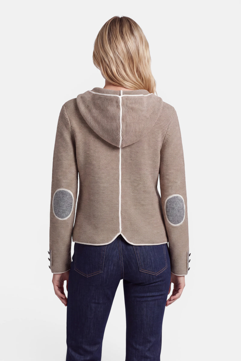 Eine Frau mit welligem blondem Haar, von hinten gesehen, trägt eine hellbraune Kapuzenjacke im bayerischen Stil mit weißem Besatz, grauen ovalen Ellbogenpatches und dunkelblauen Jeans. Die Strickjacke aus einer luxuriösen Wolle-Kaschmir-Mischung hat Knöpfe an den Ärmelbündchen und eine Mittelnaht auf der Rückseite.