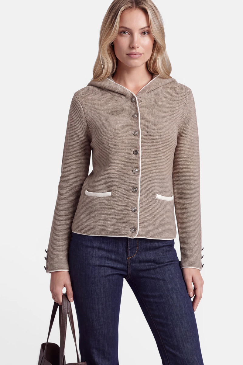 Eine Frau mit langen blonden Haaren trägt eine taupefarbene Strickjacke im bayerischen Stil mit weißem Besatz, die bis oben zugeknöpft ist und über Vordertaschen sowie Knöpfe an den Ärmeln verfügt. Dazu trägt sie eine dunkelblaue Jeans und hält eine braune Handtasche in der Hand, während sie vor einem schlichten weißen Hintergrund steht.