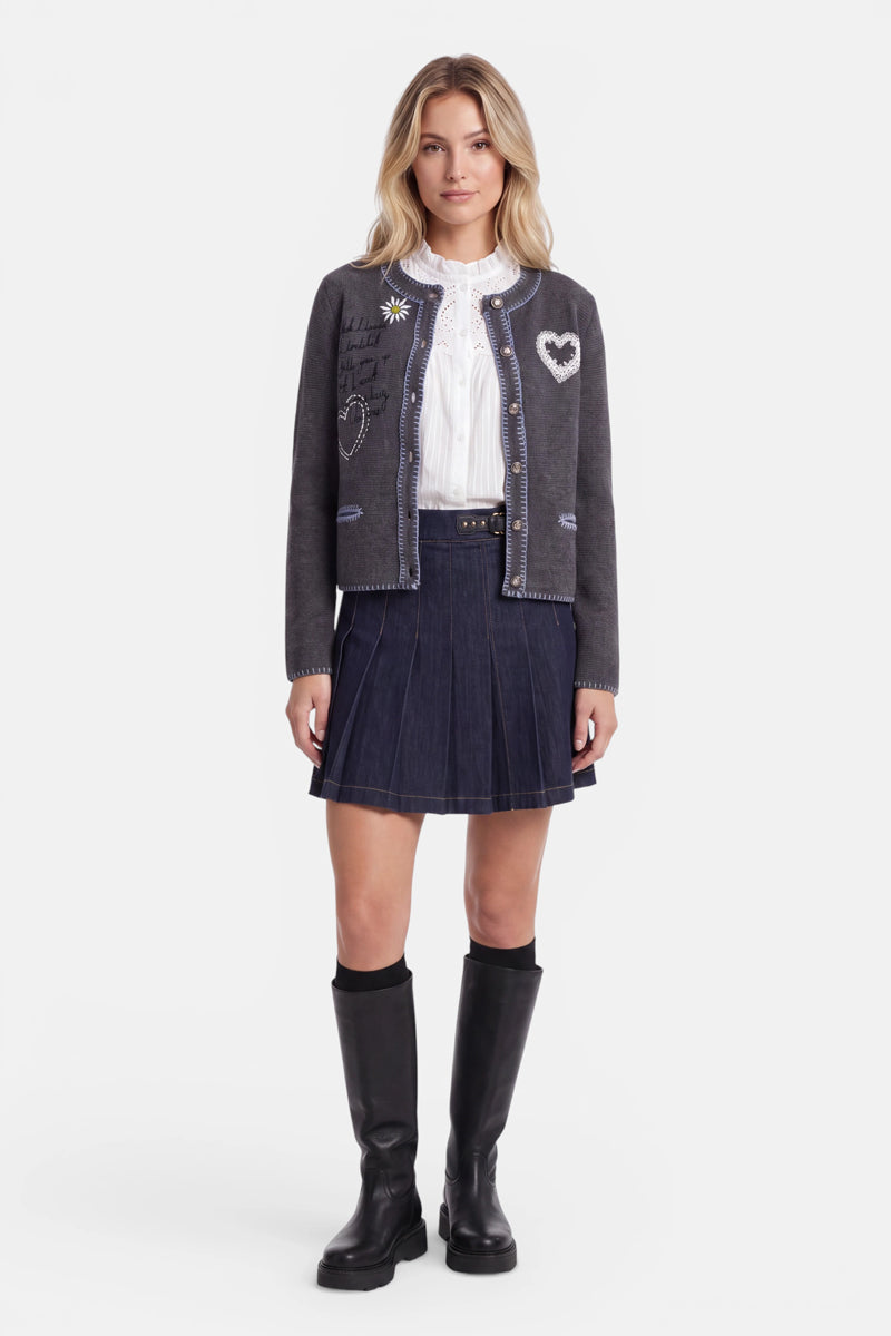 Eine Frau steht mit dem Blick nach vorne und trägt eine graue Strickjacke im bayerischen Stil, die mit zarter Herzstickerei verziert ist, eine weiße Bluse, einen marineblauen Faltenrock und schwarze kniehohe Stiefel. Sie hat helles, welliges Haar, einen neutralen Gesichtsausdruck und lässt die Arme entspannt an den Seiten hängen. Der Hintergrund ist schlicht weiß.