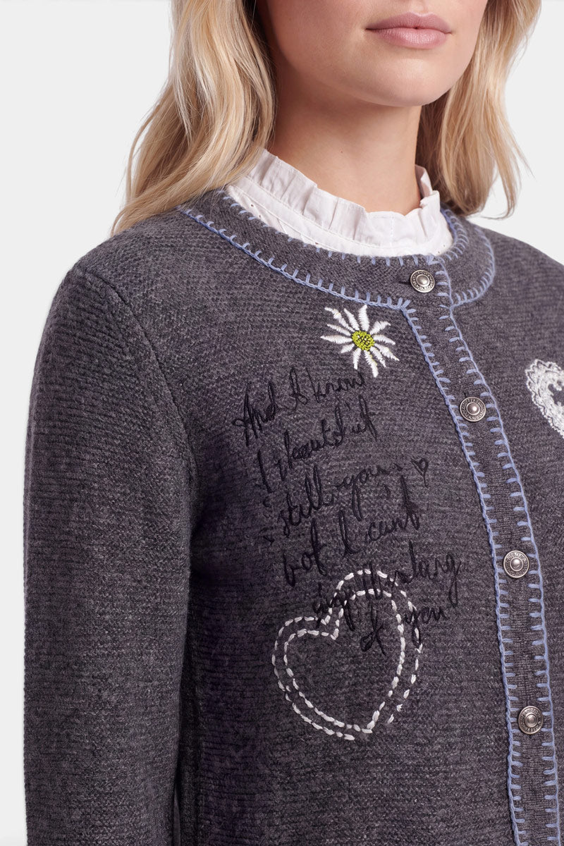 Eine Frau trägt eine graue Strickjacke im bayerischen Stil mit blauer Borte, weißer Herz- und Gänseblümchen-Stickerei sowie handgeschriebenem Text. Ihr blondes Haar trägt sie offen, und unter der Strickjacke aus einer edlen Wolle-Kaschmir-Mischung trägt sie eine weiße Bluse mit Rüschenkragen. Nur ihr Oberkörper und der untere Teil ihres Gesichts sind zu sehen.