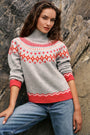 Eine Frau mit welligem braunem Haar trägt einen grauen Jacquard-Pullover mit rot-weißen Mustern im norwegischen Stil und eine blaue Jeans und steht vor einem felsigen Hintergrund. Ihre Ärmel sind hochgekrempelt, und sie blickt selbstbewusst mit einem leichten Lächeln in die Kamera.