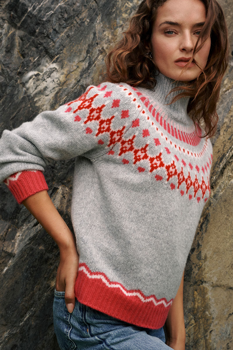 Eine Person steht im Freien vor einer Felswand und trägt einen grauen Jacquard-Pullover aus Merinowolle mit einem rot-rosa nordischen Muster an Passe und Ärmelbündchen, dazu eine blaue Jeans. Sie hat langes, welliges braunes Haar und einen neutralen Gesichtsausdruck, blickt leicht von der Kamera weg und verkörpert den klassischen norwegischen Stil.