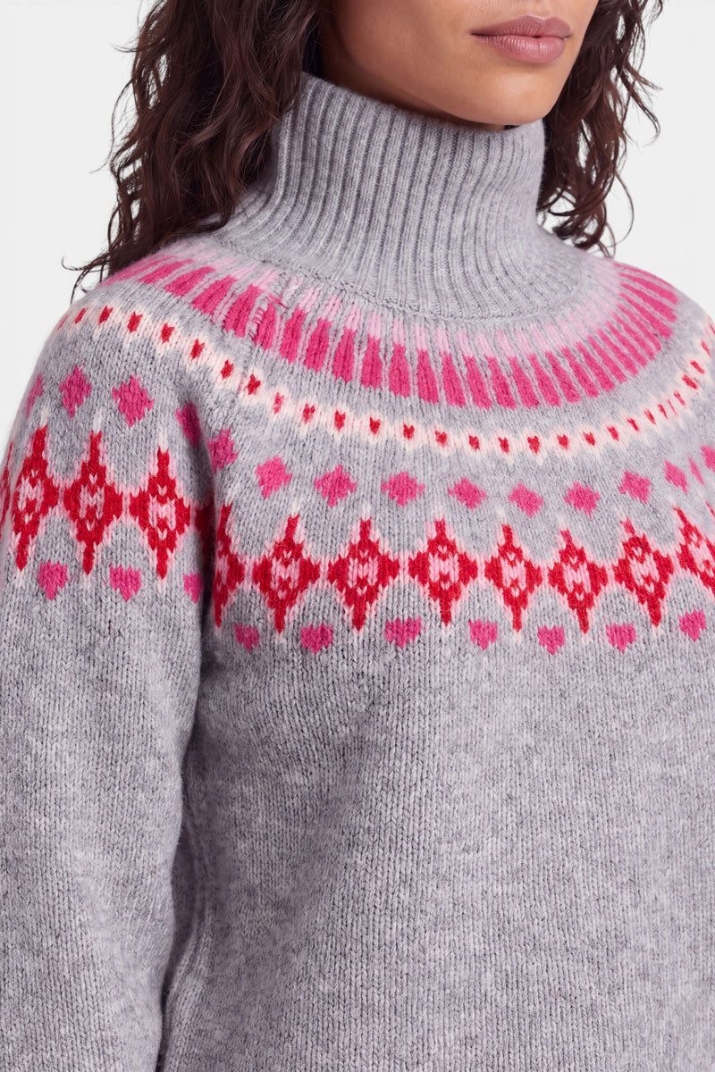 Eine Person trägt einen dicken, grauen Rollkragenpullover im norwegischen Stil mit einem rosa-roten Muster aus geometrischen Formen und Herzen auf Brust und Schultern. Die Person hat welliges braunes Haar, und ihr Gesicht ist teilweise zu sehen. Der Hintergrund ist einfarbig weiß.