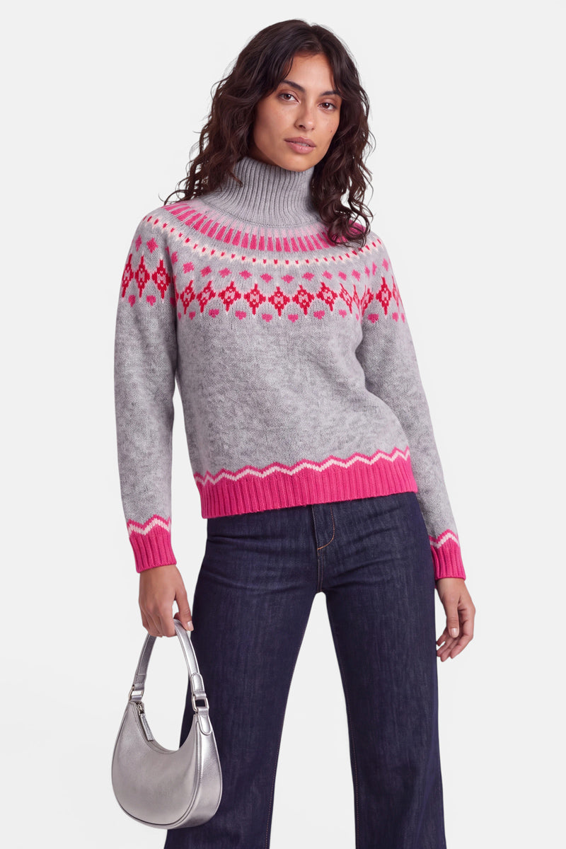 Eine Frau mit gewelltem Haar trägt einen grauen Rollkragenpullover im norwegischen Stil mit rosa und roten geometrischen Mustern, dazu eine dunkle Jeans. Sie hält eine silberne Handtasche an ihrer Seite und steht vor einem schlichten weißen Hintergrund, wobei sie selbstbewusst in die Kamera blickt.