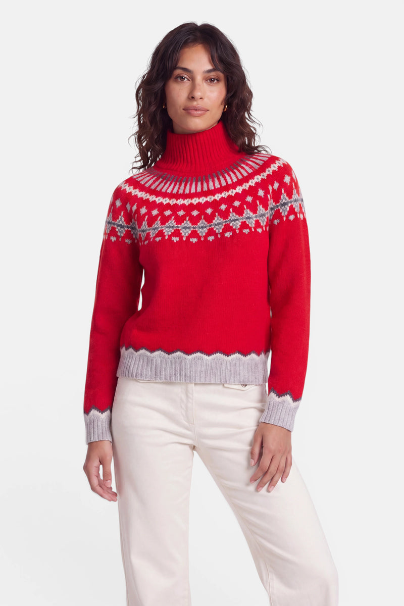 Eine Frau mit welligem, dunklem Haar trägt einen roten Jacquard-Pullover im Norweger-Stil mit einem grau-weißen geometrischen Muster und grauen Bündchen. Dazu trägt sie eine cremefarbene Hose. Der Hintergrund ist schlicht weiß, und sie blickt mit entspannter Haltung selbstbewusst in die Kamera.