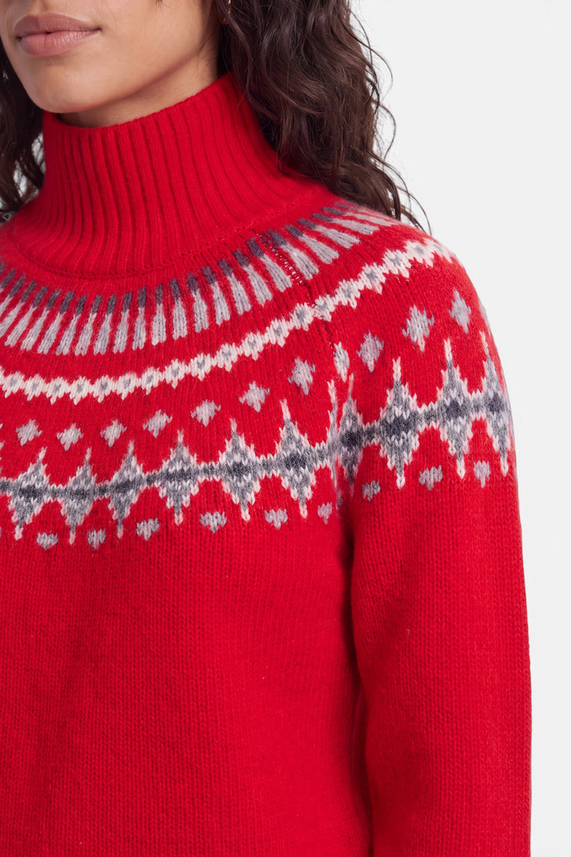 Eine Person, die einen leuchtend roten Rollkragenpullover im norwegischen Stil trägt, mit einem grau-weißen geometrischen Fair-Isle-Muster über Brust und Schultern; nur der untere Teil des Gesichts, der Hals und der obere Oberkörper sind zu sehen.
