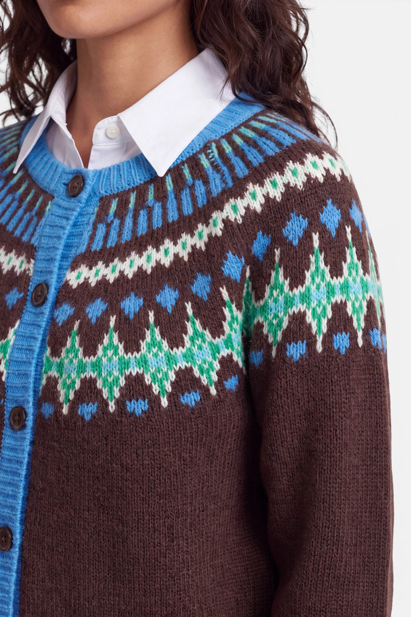 Eine Frau, die einen braunen Strickpullover im norwegischen Stil mit blauen, grünen und weißen geometrischen Mustern über einem weißen Hemd mit Kragen trägt. Die Jacquard-Strickjacke hat blaue Rippbündchen und dunkle Knöpfe. Zu sehen sind nur die untere Hälfte ihres Gesichts und ihr Oberkörper.