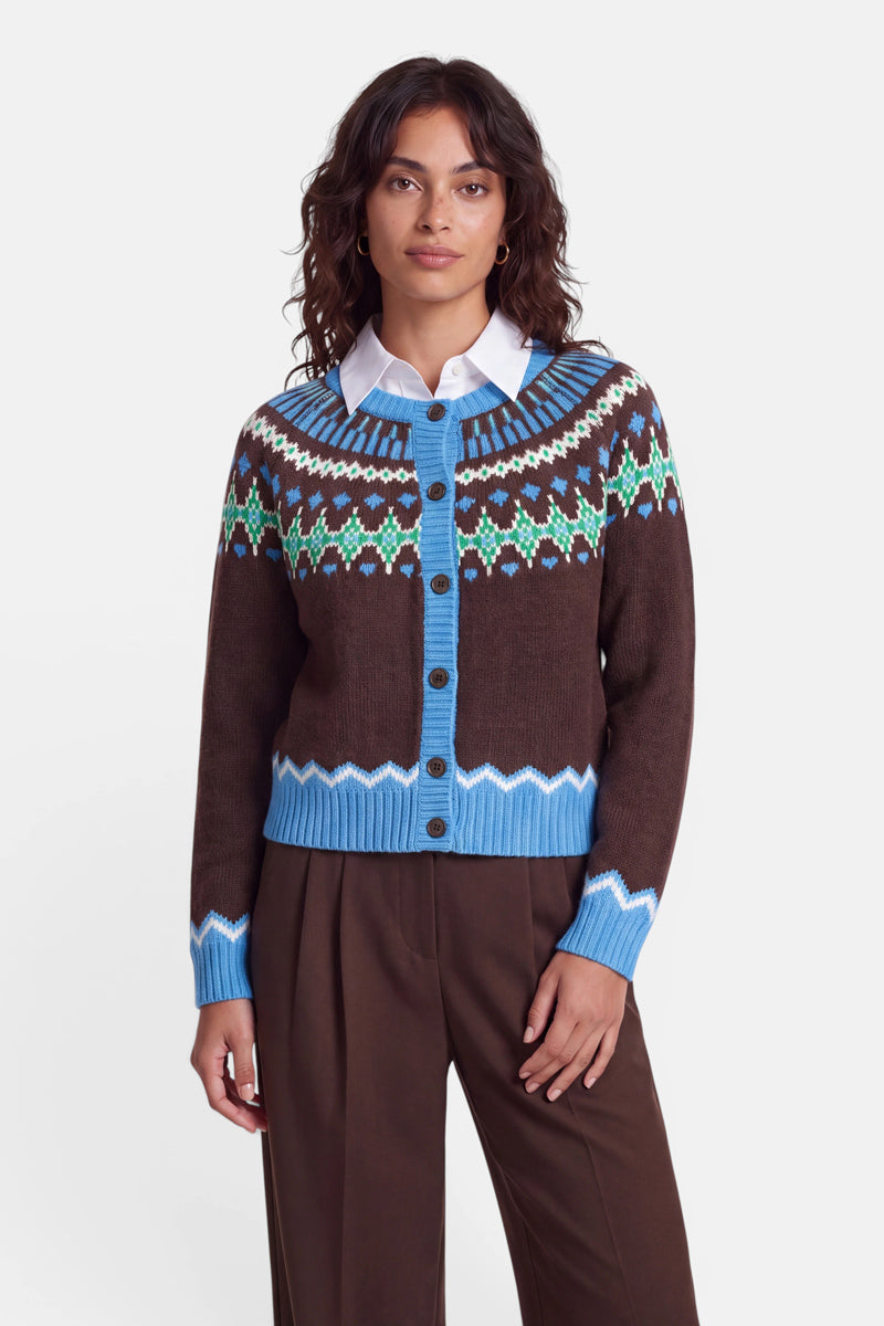 Eine Frau mit welligem, dunklem Haar trägt eine braune Jacquard-Strickjacke mit blauen und grünen geometrischen Mustern im norwegischen Stil über einem weißen Hemd mit Kragen. Die Strickjacke hat einen blauen, gerippten Saum und Ärmelbündchen. Außerdem trägt sie eine braune Hose und steht vor einem weißen Hintergrund.