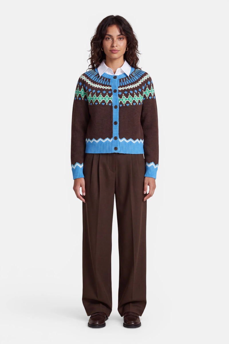 Eine Frau steht mit dem Blick nach vorne und trägt eine Jacquard-Strickjacke im Norweger-Stil mit blauen, grünen und weißen Mustern über einem hellblauen Hemd mit Kragen. Sie hat offenes dunkles Haar und trägt eine weite braune Hose sowie dunkle Schuhe; sie steht vor einem schlichten weißen Hintergrund.