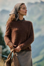 Eine Frau, die einen flauschigen Pullover in sattem Braun und einen grauen Rock trägt, steht im Freien vor einer Bergkulisse. Sie hat einen grauen Schal über dem Haar und hält eine braune Tasche in der Hand. Ihr Gesichtsausdruck ist ruhig, und die Szene ist sanft beleuchtet und strahlt eine natürliche, friedliche Atmosphäre aus.