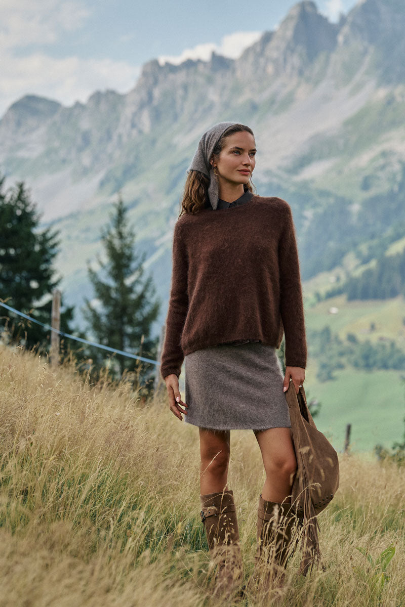 Eine Frau steht auf einer grasbewachsenen Wiese, im Hintergrund sind Berge zu sehen. Sie trägt einen flauschigen Pullover, einen grauen Rock, braune Stiefel und ein Kopftuch. Sie hält eine braune Tasche in der Hand und blickt in die Ferne. Hinter ihr sind Kiefern und ein blauer Himmel zu sehen.