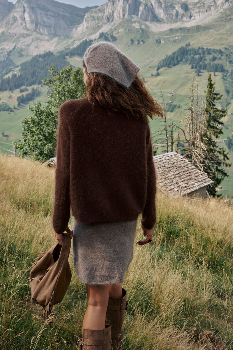 Eine Person in einem braunen Pullover aus gebürsteter Wolle, einem grauen Rock und einer grauen Baskenmütze geht durch hohes Gras auf eine rustikale Hütte zu, im Hintergrund sind in der Ferne Berge und Wälder zu sehen. Sie trägt eine braune Tasche und kniehohe braune Stiefel, um die Schultern ist ein flauschiger Schal drapiert. Die Szene wirkt ruhig und natürlich.