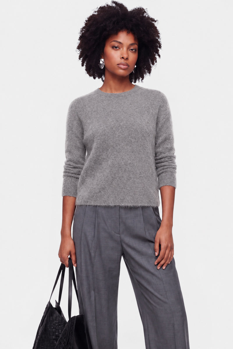 Eine Frau mit natürlich lockigem Haar trägt einen flauschigen Pullover aus gebürsteter Wolle mit Rundhalsausschnitt und eine lockere graue Hose. Sie steht vor einem schlichten weißen Hintergrund, hält eine schwarze, strukturierte Tragetasche in der Hand und blickt mit ausdruckslosem Gesicht geradeaus.