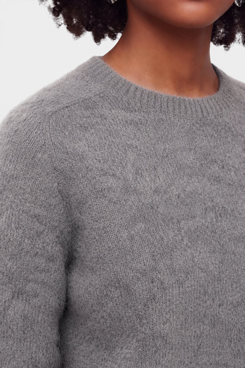 Eine Person mit mittelbrauner Haut und lockigem Haar trägt einen weichen, strukturierten grauen Pullover mit Rundhalsausschnitt aus gebürsteter Wolle. Vor einem schlichten weißen Hintergrund sind nur der untere Teil des Gesichts, der Hals und der obere Oberkörper zu sehen.